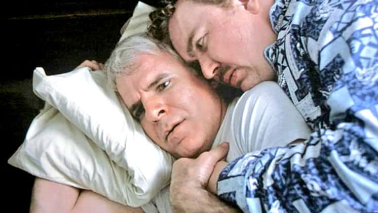 Del, portrayed by John Candy is cuddling Neal, portrayed by Steve Martin are stuck in a crappy motel, forced to share a bed. In this image, they wake up, discovering they had been cuddling all night.