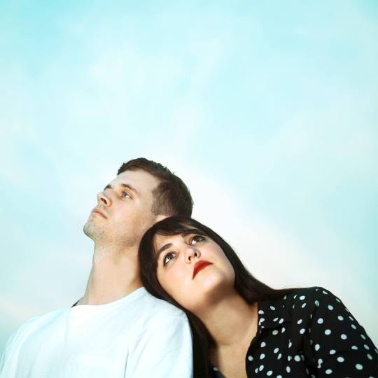 Against a blue background, Allie McDonald rests her head on the shoulder of Mike Derenzo and they stare up into the distance.