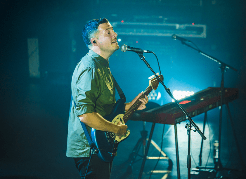 SYML, wearing a green button up shirt, plays his blue and white electric guitar and sings intro the microphone. 