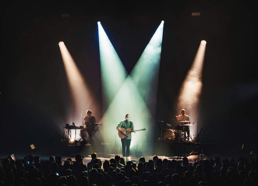 SYML stands on stage, playing an acoustic guitar. He is illuminated by two spotlights that originate behind him. Behind him on his left and right are two of his band members, one playing piano and the other sitting at a drum set. 