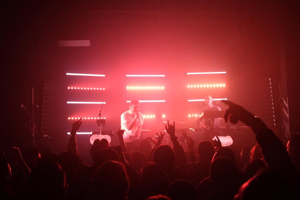 The duo that is RKCB preforms on stage, back lit by red lights arranged in horizontal lines. Members of the crowd have their hands up. 