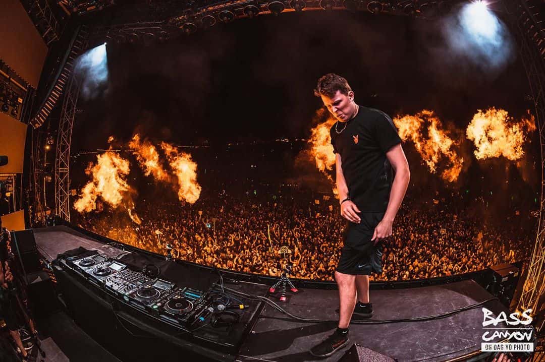 UBUR on top of the DJ Table in front of a large crowd at the Gorge Amphitheater for Bass Canyon 2019 while pyrotechnics/flame cannons go off in front of the stage.