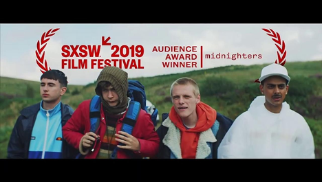 Four high school aged boys are looking towards the camera, but off into the distance. They are wearing multiple layers of warm fall jackets. Behind them is a green grass hill and a cloudy sky.