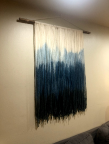 many pieces of yarn that are tied across a wooden dowel. The yarn is dyed an ombre blue. It is hanging from the wall by a piece of rope tied from the edges of the dowel to one hook.