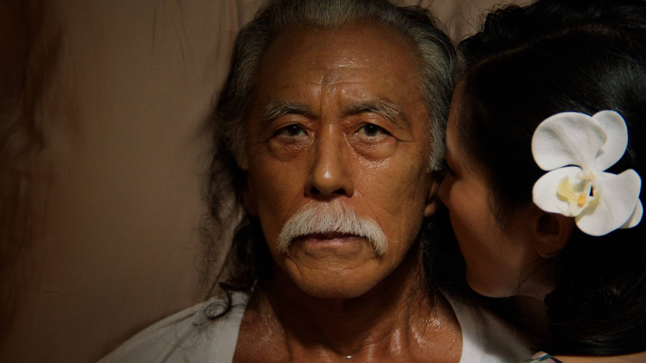 I Was A Simple Man STILL picturing an old man, the family Patriarch Masao Matsuyoshi (Steve Iwamoto). He has a blank expression but looks to be sweating as the ghost of his wife Grace (Constance Wu) is whispering in his ear, a large flower in her hair
