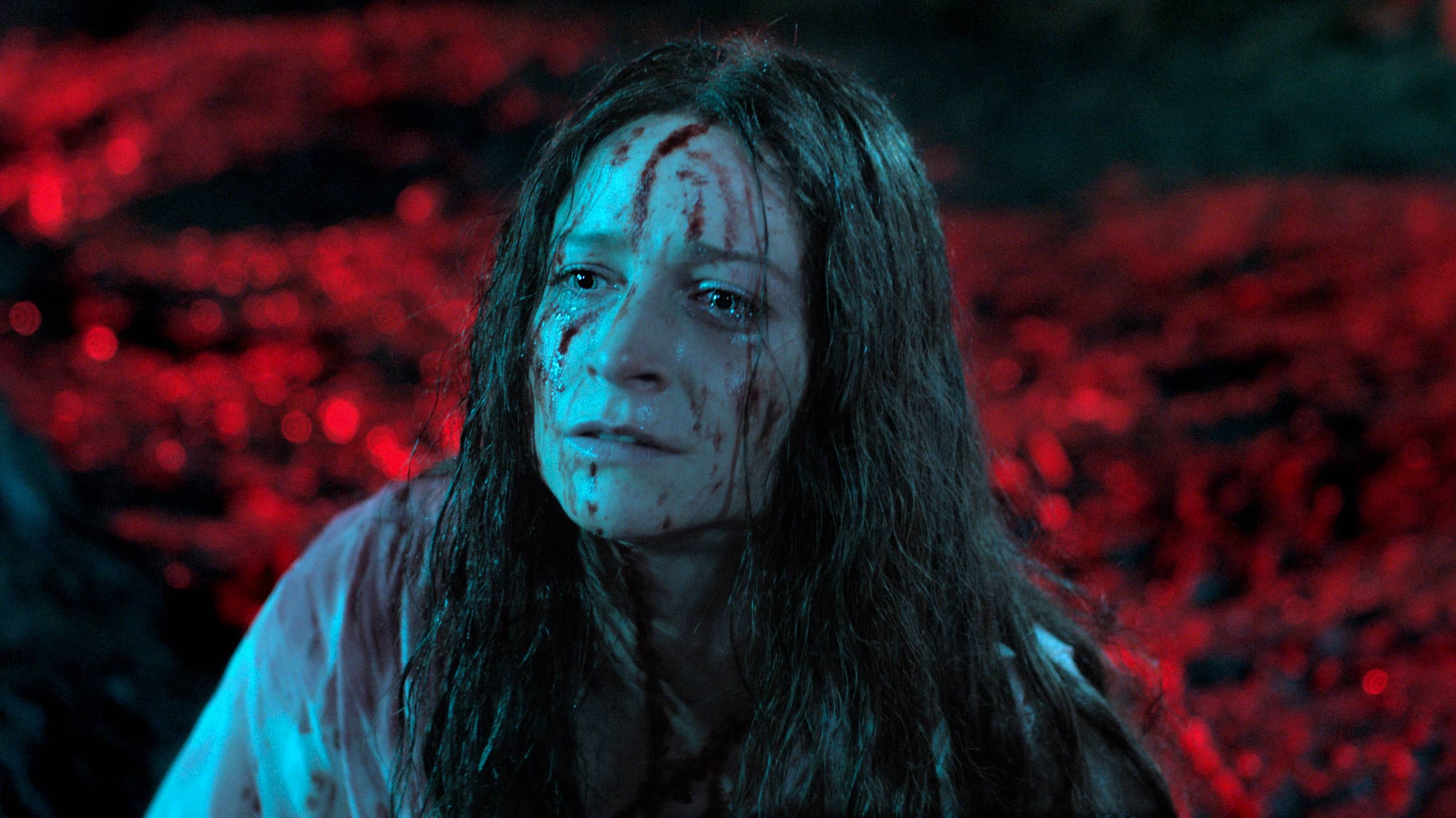 A picture of Enid (Niamh Algar) in her “final girl” scene. The background is a blurry red, focused only on disheveled Enid, who is covered in blood and tears. There is blood matting her hair and blood contrasting on her white dress.