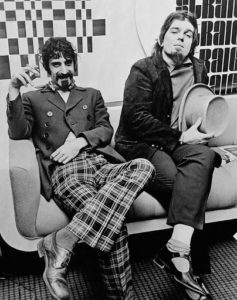 Frank Zappa is seated on a couch smoking a cigarette. Sitting to his left is Captain Beefheart. They both have their legs crossed and are dressed in suits.