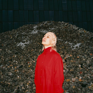 Cate le Bon looks out of frame. She is wearing a red blouse and standing in front of a large pile of small rocks.