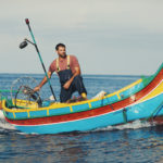 Jesmark Scicluna steers a small blue, red, and yellow fishing boat (known as a luzzu) in the open ocean.