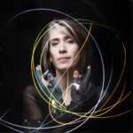 Imogen Heap at the Moody Theater 2019. She is standing with her hand crossed in front of her face as light trails flow around her.