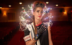 Imogen Heap is standing in a theater, with the seats behind her. She is wearing a black top with an arc of lights attached to her shoulders that moves behind her head. 