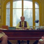 An aged Bill Murray sits in the center of the image behind a dark wood desk in a yellow office. In the left part of the image is Angellica Bette Fellini who is sitting on top of a desk, and sitting in a chair on the right side of the image is Elizabeth Moss.