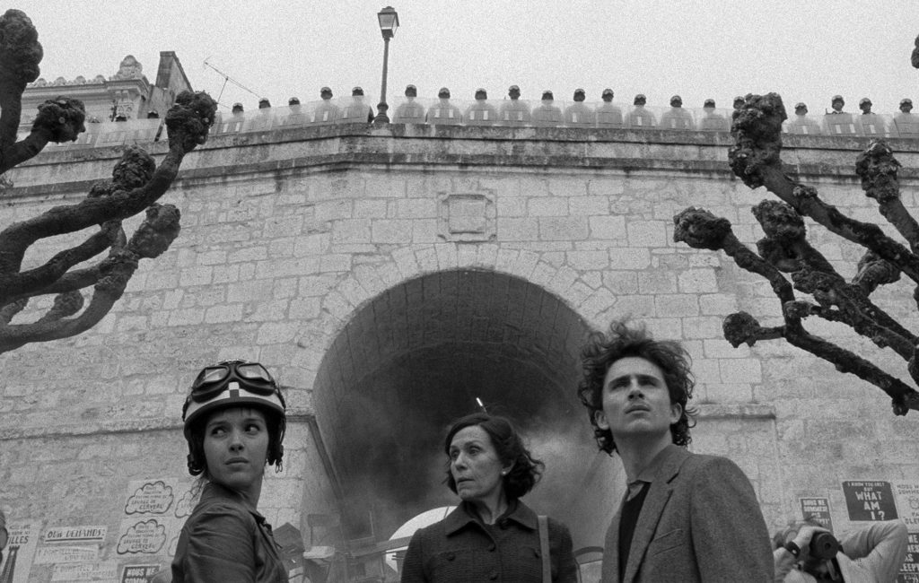From left to right, Lyna Khoudri, Frances McDormand, and Timothée Chalamet stand below a large stone wall. On top of the wall is a row of police officers in riot gear. The image is in black and white.