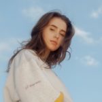 photo of Clairo, in a white shirt with a blue sky with clouds in the background