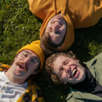 Three members of Diet Lite are laying on their backs on the grass with their heads touching each other in unision