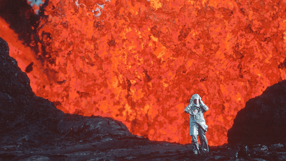 A scientist stands beneath a towering eruption of bright red lava in a silver protective suit.