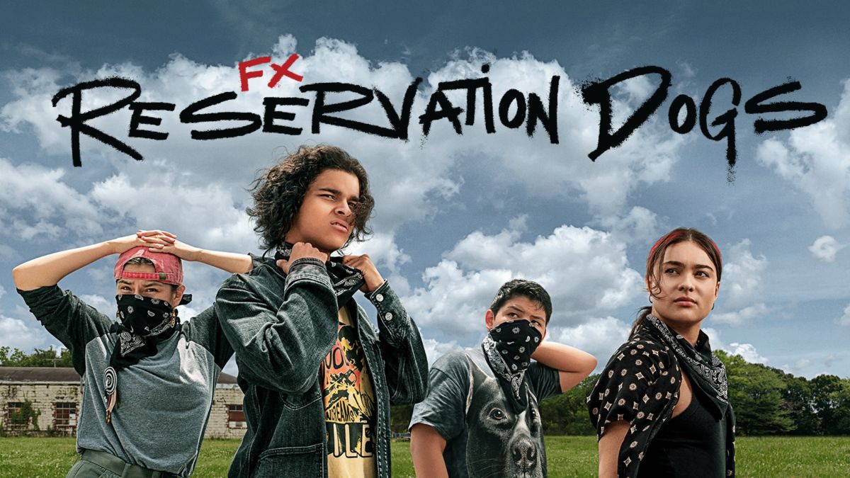 Two teenage girls and two teenage boys dressed in dark clothing with bandanas around their necks are standing outside. The title “Reservation Dogs” in black, smudged handwriting is written above them.